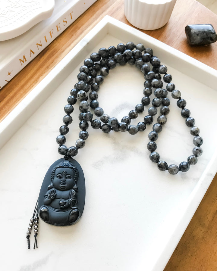 A 108 beaded mala necklace with Larvikite beads and a Black Obsidian Buddha pendant, displayed on a white dish alongside a manifestation diary and small crystals.
