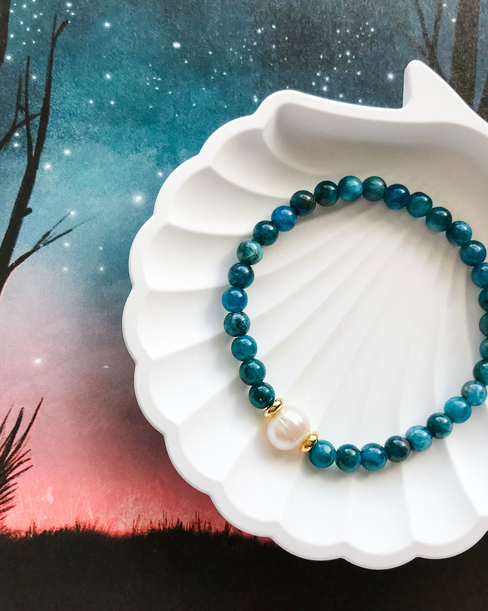 A mala bracelet with blue Apatite beads and a freshwater pearl centerpiece, displayed on a white dish against a stylized ocean-themed background.
