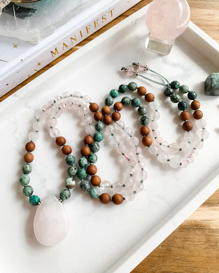 A hand-knotted mala necklace with beads of African Turquoise, Rose Quartz, and Sandalwood, and silver accents, with a Rose Quartz teardrop shaped pendant, displayed on a marble tray with small crystals.