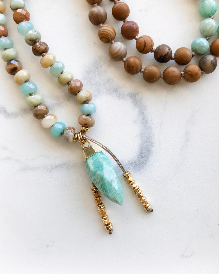 A mala necklace with a combination of blue-green Amazonite, brown Matte Petrified Wood, and beige Picture Jasper beads, displayed on a white surface with a gold-dipped Amazonite pendant.