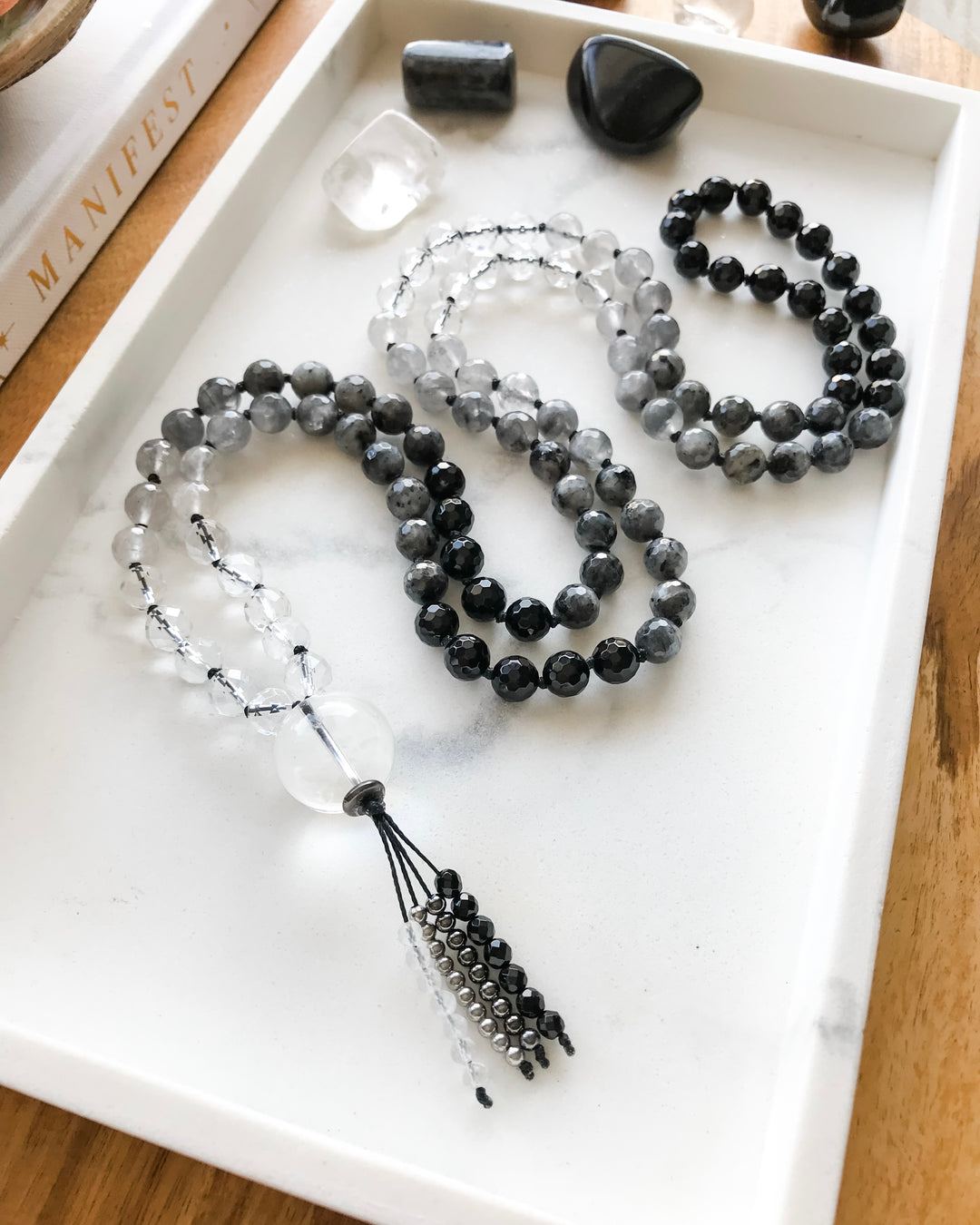 A mala necklace with black onyx, clear quartz, grey quartz and larvikite beads and clear quartz guru bead, displayed on a white tray with crystals and a book.