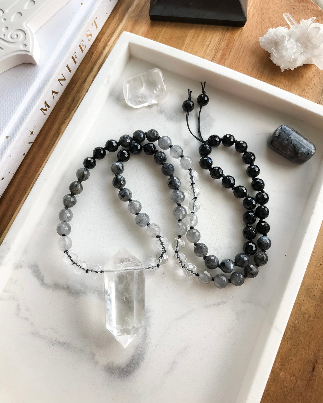A short mala necklace laid out on a white surface, consisting of black onyx, clear quartz, grey quartz, and larvikite beads, with a clear quartz pendant, surrounded by small crystals.
