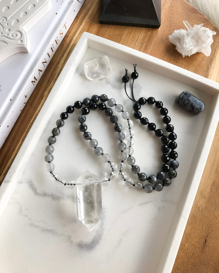 A short mala necklace laid out on a white surface, consisting of black onyx, clear quartz, grey quartz, and larvikite beads, with a clear quartz pendant.