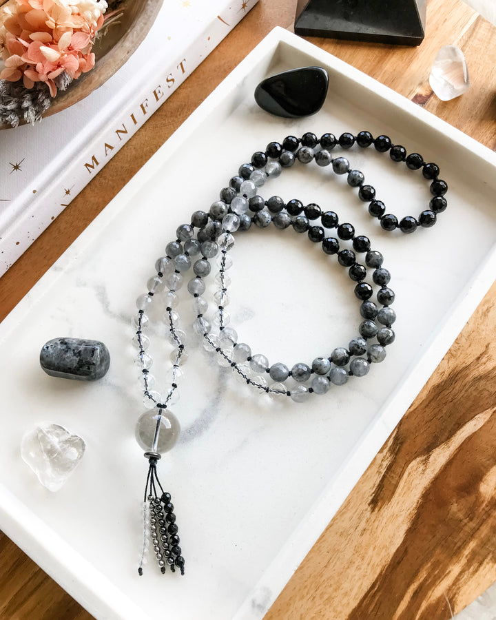 A mala necklace with black onyx, clear quartz, grey quartz and larvikite beads and clear quartz guru bead, displayed on a white tray with crystals and a book.
