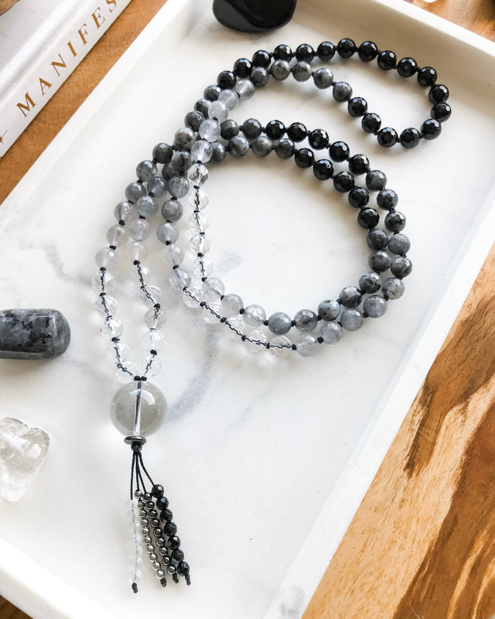 A mala necklace with black onyx, clear quartz, grey quartz and larvikite beads and clear quartz guru bead, displayed on a white tray with crystals and a book.