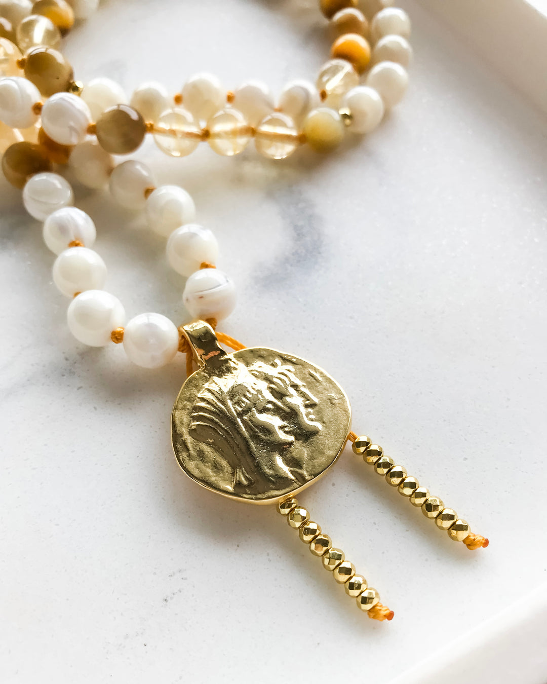 A mala necklace with Mother of Pearl, Citrine, and Honey Tiger Eye beads, featuring gold-colored accents and a gold-tone pendant, displayed on a marble surface.