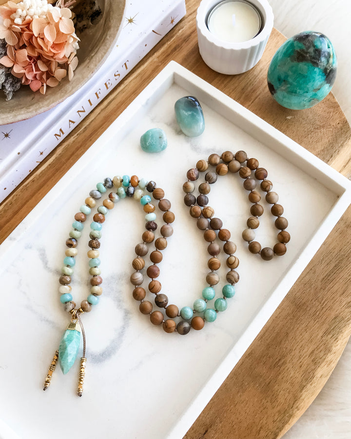 A mala necklace with a combination of blue-green Amazonite, brown Matte Petrified Wood, and beige Picture Jasper beads, displayed on a white surface with a gold-dipped Amazonite pendant, surrounded by crystals and a flower smudge stick.