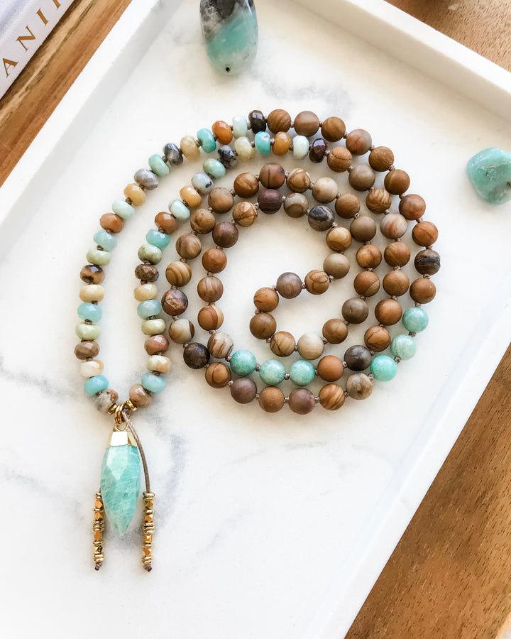 A mala necklace with a combination of blue-green Amazonite, brown Matte Petrified Wood, and beige Picture Jasper beads, displayed on a white surface with a gold-dipped Amazonite pendant.