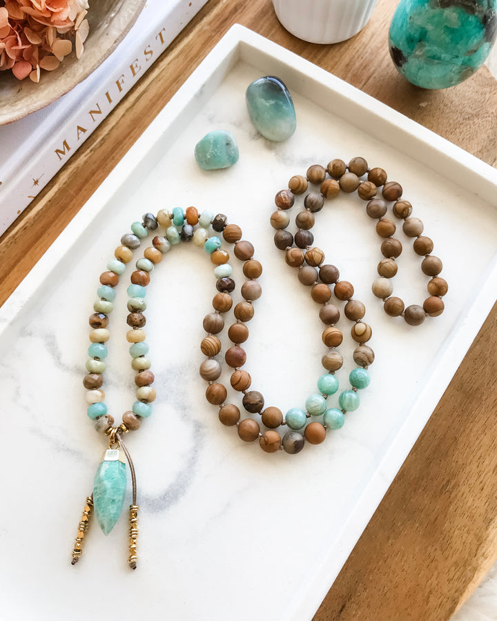 A mala necklace with a combination of blue-green Amazonite, brown Matte Petrified Wood, and beige Picture Jasper beads, displayed on a white surface with a gold-dipped Amazonite pendant, surrounded by crystals and a flower smudge stick.