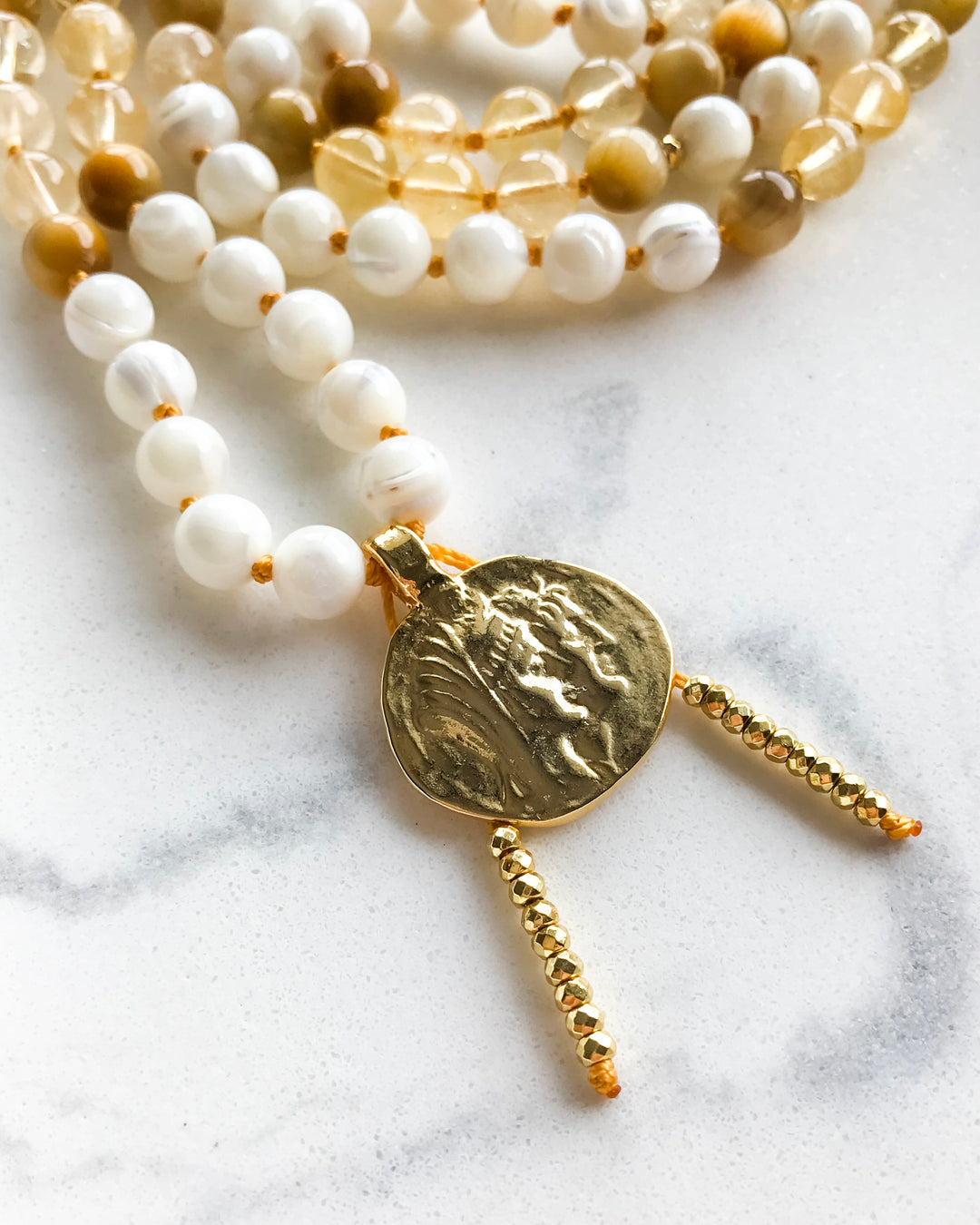 A mala necklace with Mother of Pearl, Citrine, and Honey Tiger Eye beads, featuring gold-colored accents and a gold-tone pendant, displayed on a marble surface.