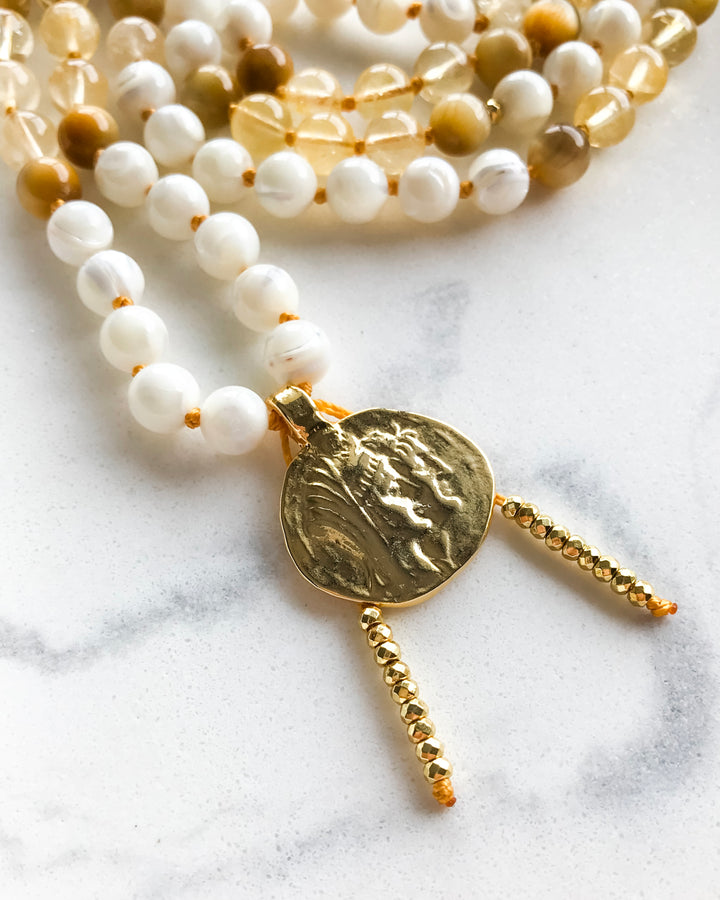 A mala necklace with Mother of Pearl, Citrine, and Honey Tiger Eye beads, featuring gold-colored accents and a gold-tone pendant, displayed on a marble surface.