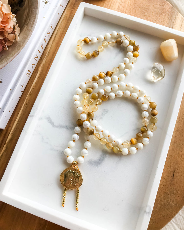 A mala necklace with Mother of Pearl, Citrine, and Honey Tiger Eye beads, featuring gold-colored accents and a gold-tone pendant, displayed on a marble surface surrounded by small crystals.