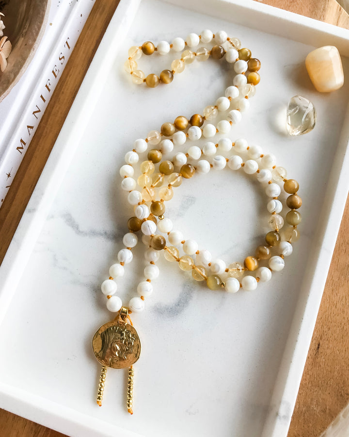 A mala necklace with Mother of Pearl, Citrine, and Honey Tiger Eye beads, featuring gold-colored accents and a gold-tone pendant, displayed on a marble surface.
