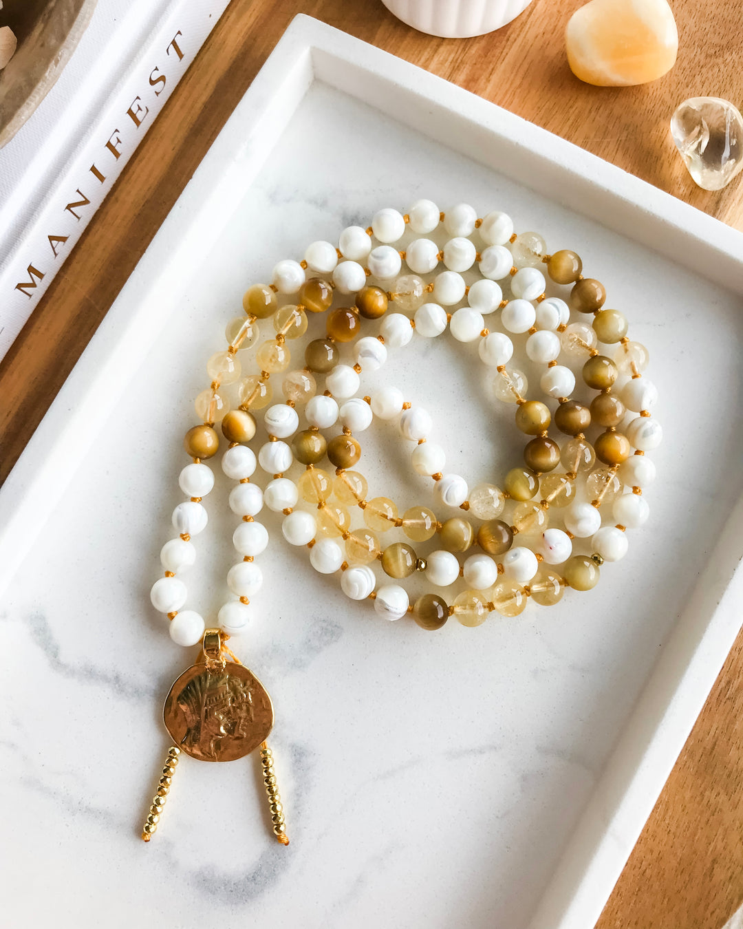 A mala necklace with Mother of Pearl, Citrine, and Honey Tiger Eye beads, featuring gold-colored accents and a gold-tone pendant, displayed on a marble surface surrounded by small crystals.