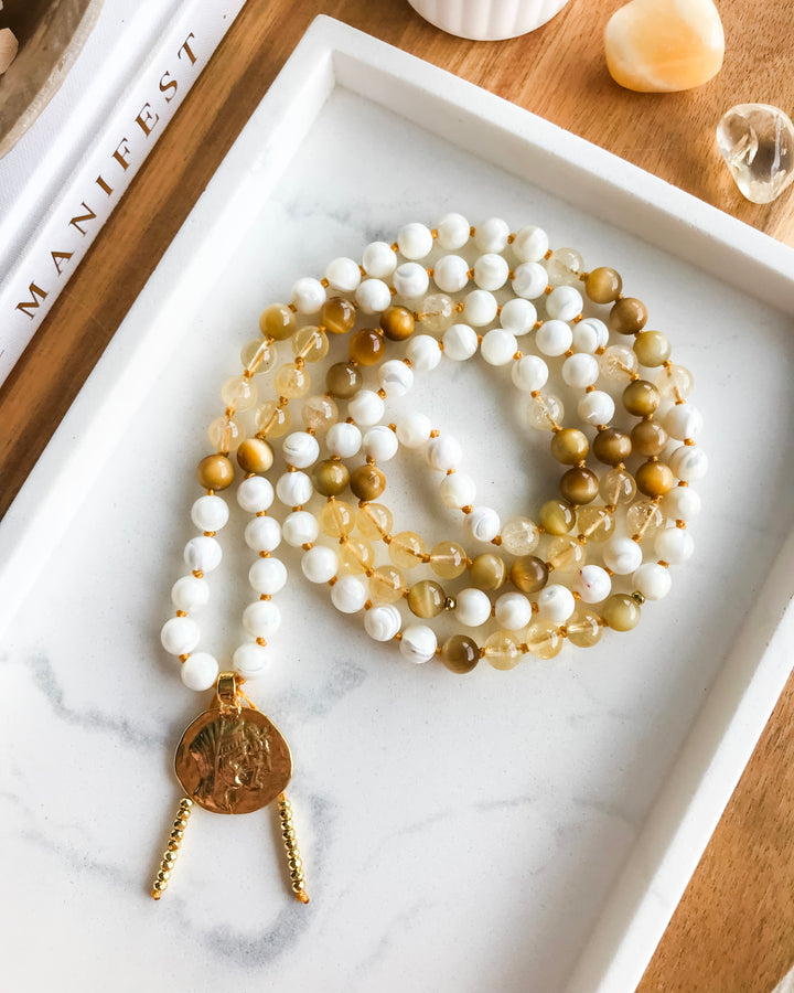 A mala necklace with Mother of Pearl, Citrine, and Honey Tiger Eye beads, featuring gold-colored accents and a gold-tone pendant, displayed on a marble surface surrounded by small crystals.