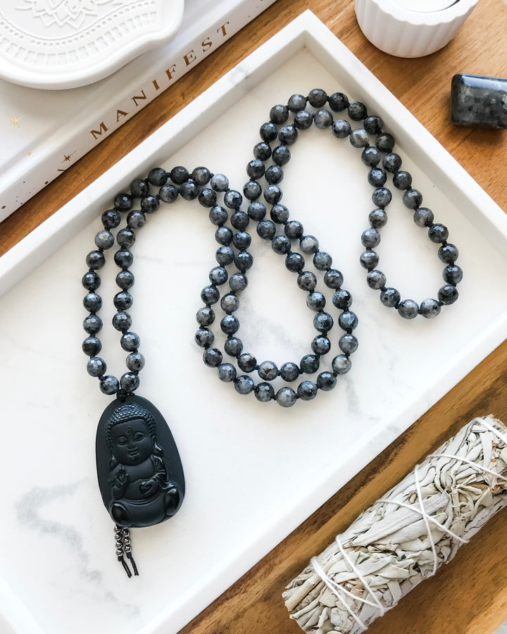 A 108 beaded mala necklace with Larvikite beads and a Black Obsidian Buddha pendant, displayed on a white dish alongside a smudge stick and small crystals.