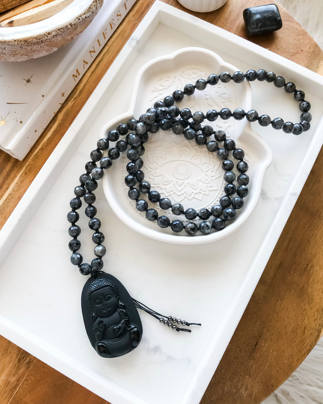 A 108 beaded mala necklace with Larvikite beads and a Black Obsidian Buddha pendant, displayed on a white dish alongside small crystals.