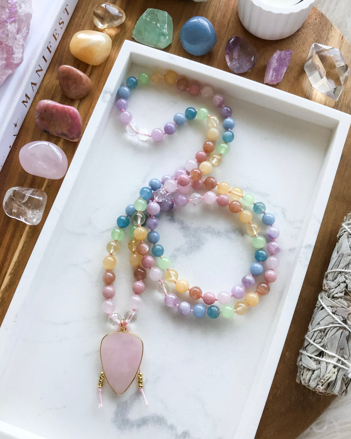 A mala necklace with a variety of colorful beads, knotted and finished with a rose quartz pendant, displayed on a white dish alongside other crystals and a smudge stick.