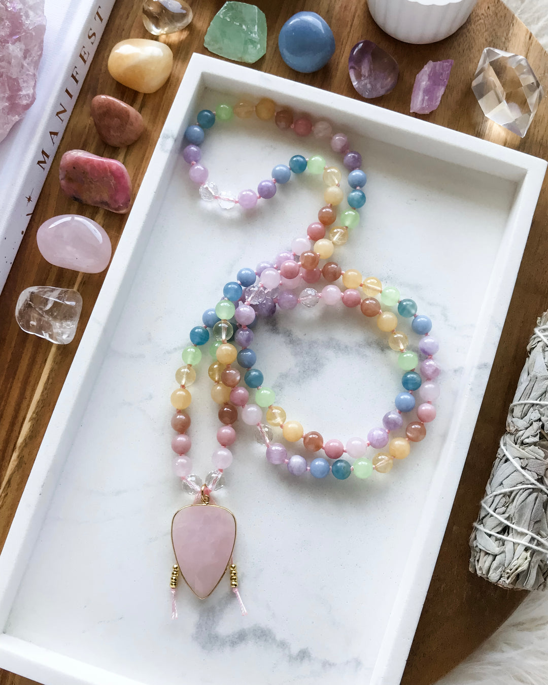 A mala necklace with a variety of colorful beads, knotted and finished with a rose quartz pendant, displayed on a white dish alongside other crystals and a smudge stick.