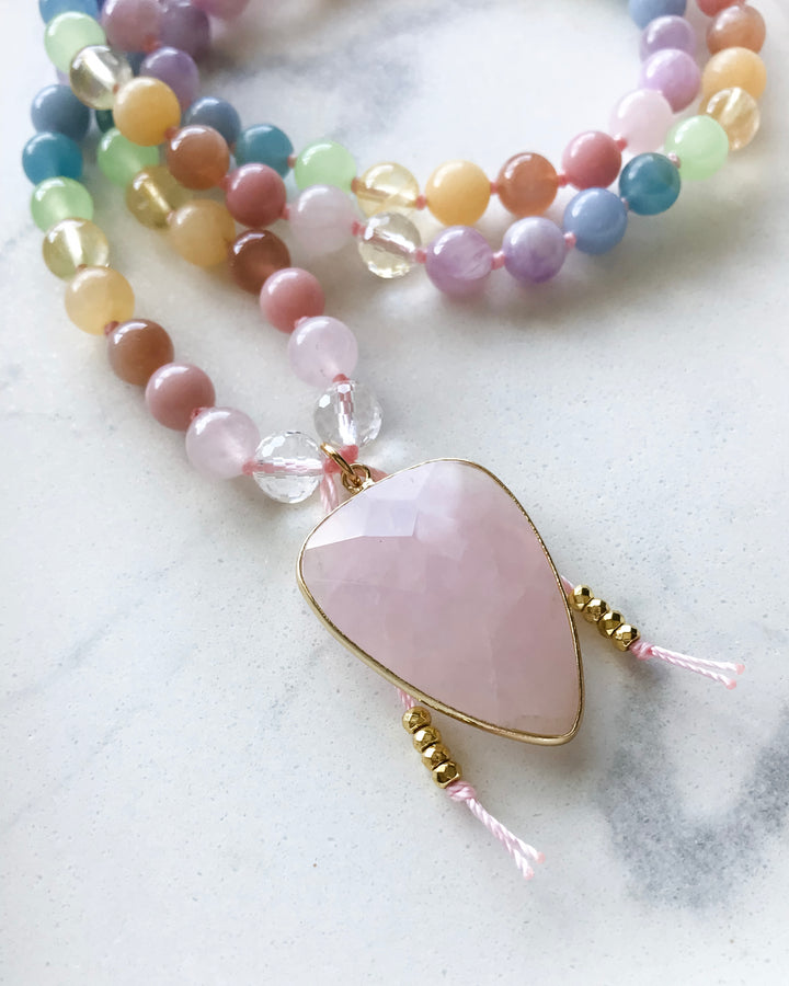 A mala necklace with a variety of colorful beads, knotted and finished with a rose quartz pendant, displayed on a white dish alongside other crystals and a smudge stick.