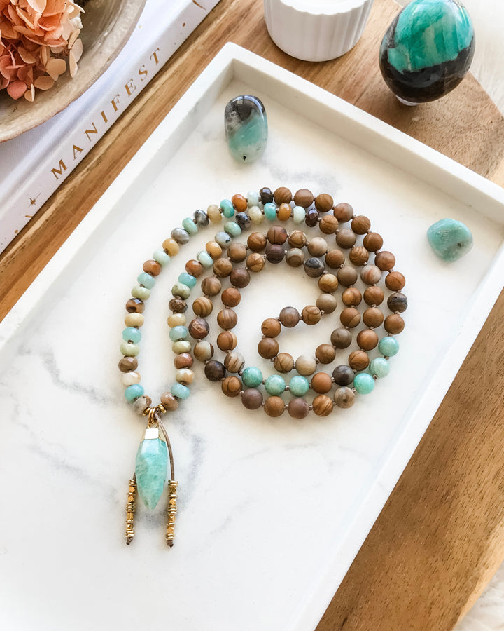 A mala necklace with a combination of blue-green Amazonite, brown Matte Petrified Wood, and beige Picture Jasper beads, displayed on a white surface with a gold-dipped Amazonite pendant, surrounded by crystals and a flower smudge stick.