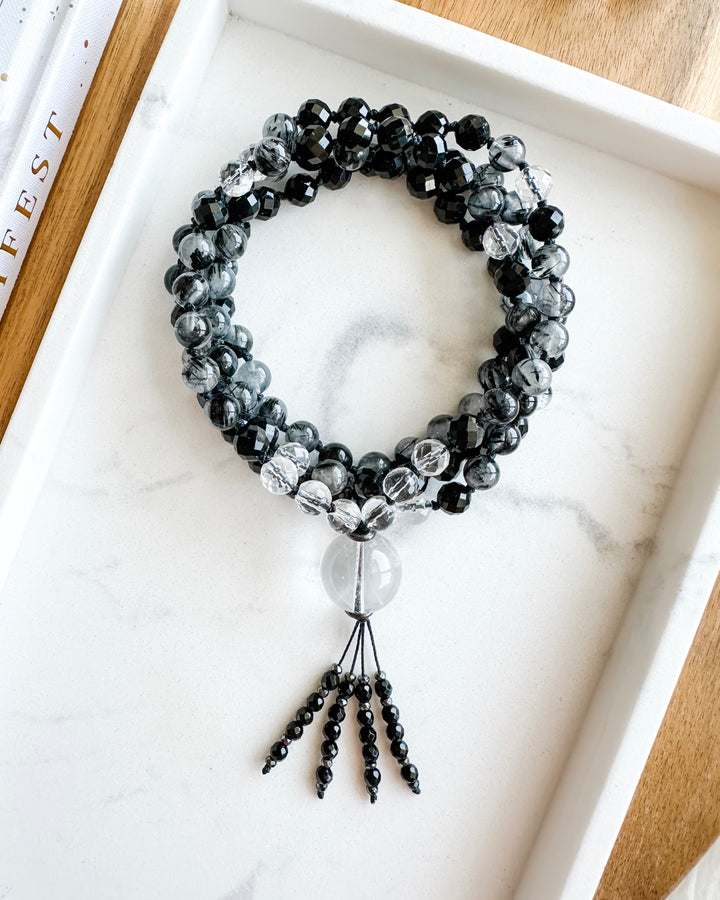 A mala necklace with black tourmaline, clear quartz, tourmalinated quartz, and hematite beads, hand-knotted and displayed on a marble tray.
