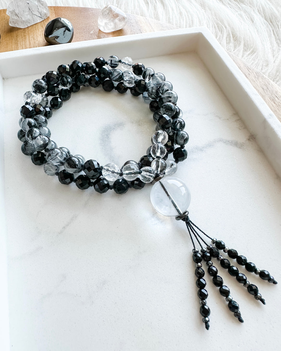 A mala necklace with black tourmaline, clear quartz, tourmalinated quartz, and hematite beads, hand-knotted and displayed on a marble tray, surrounded by small crystals.
