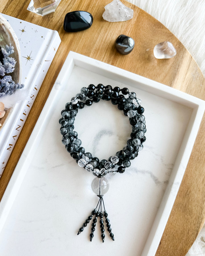 A mala necklace with black tourmaline, clear quartz, tourmalinated quartz, and hematite beads, hand-knotted and displayed on a marble tray, surrounded by small crystals and a book.