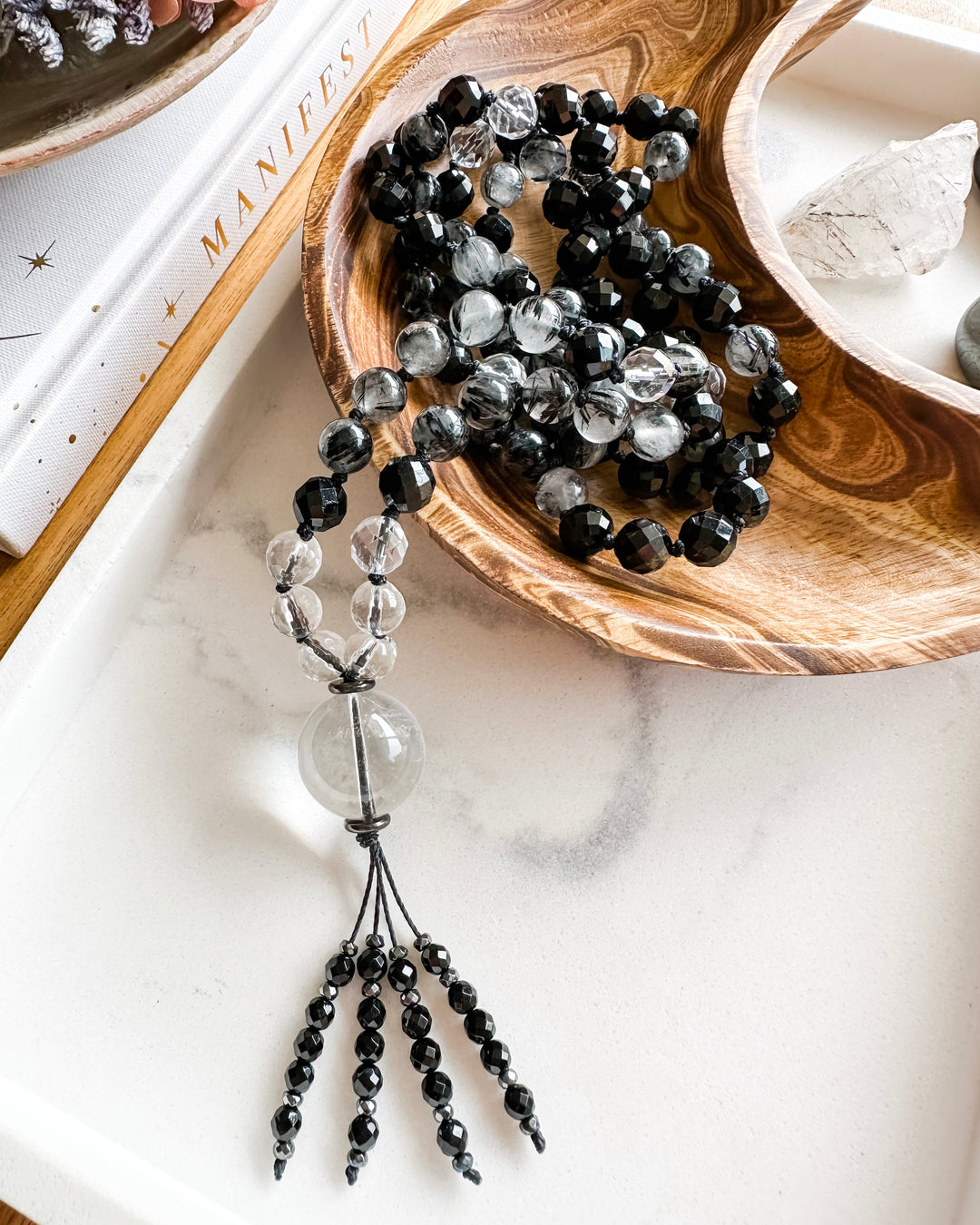 A mala necklace with black tourmaline, clear quartz, tourmalinated quartz, and hematite beads, hand-knotted and displayed on a marble tray, surrounded by small crystals and a book.