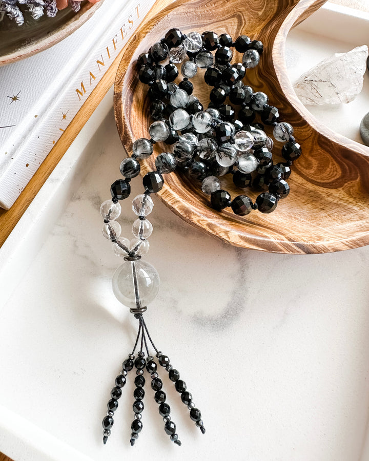 A mala necklace with black tourmaline, clear quartz, tourmalinated quartz, and hematite beads, hand-knotted and displayed on a marble tray, surrounded by small crystals and a book.