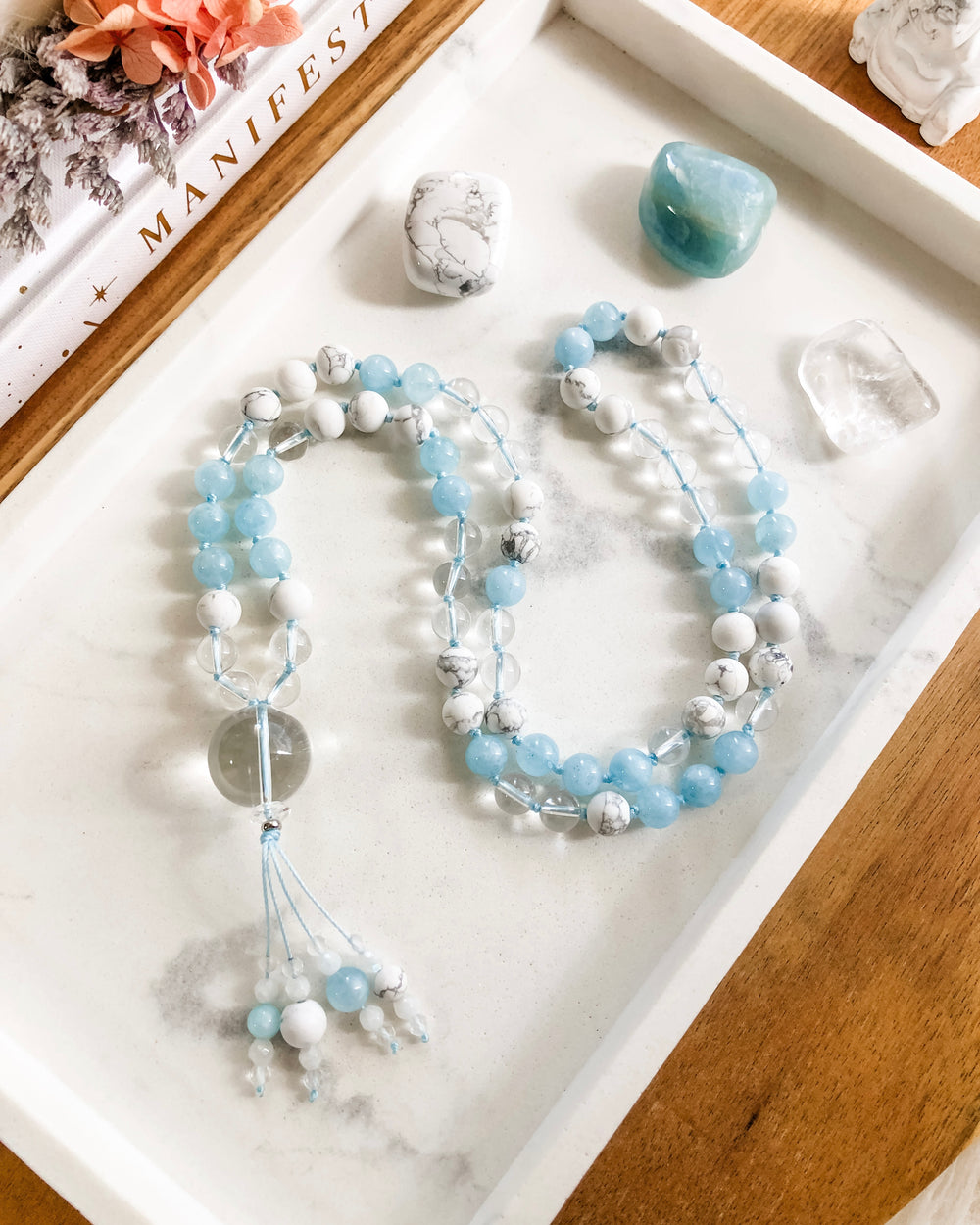 A hand-knotted mala necklace with Aquamarine, Clear Quartz, and White Howlite beads, featuring a Clear Quartz pendant and a tassel with various beads, placed on a marble tray.
