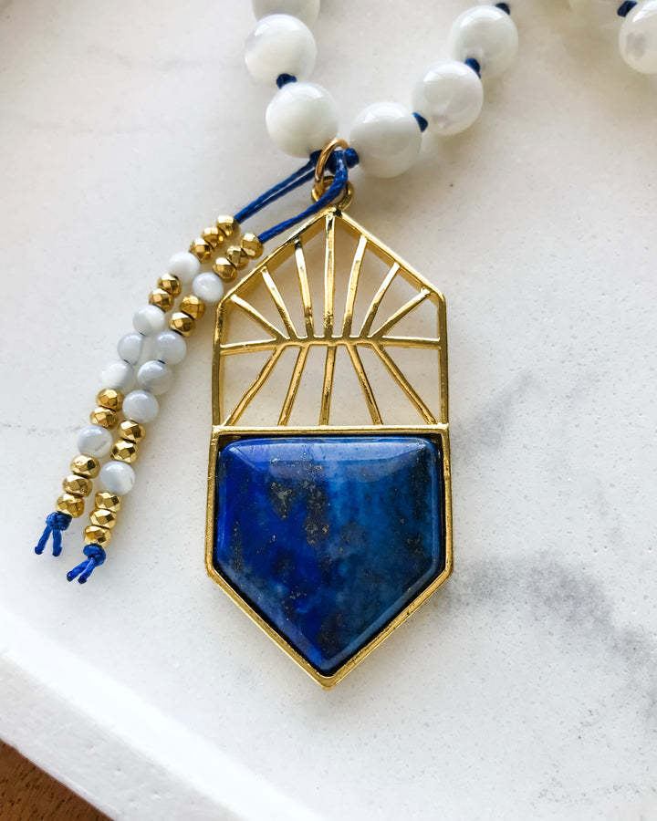 A mala necklace with Mother of Pearl beads and Lapis Lazuli accents, featuring a Lapis Lazuli pendant, displayed on a white tray.