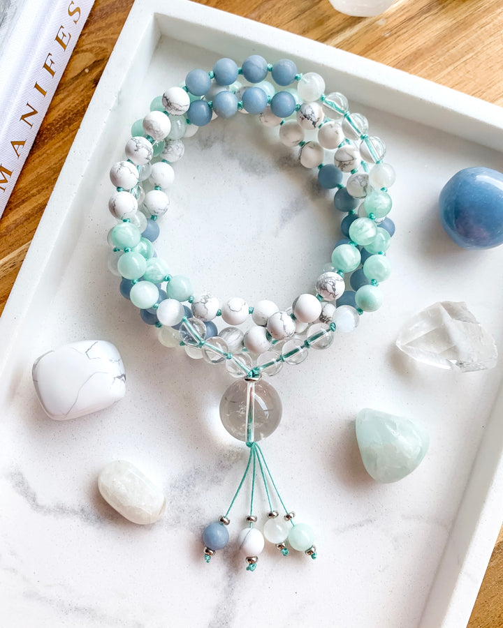 A hand-knotted mala necklace with Clear Quartz, Selenite, Green Moonstone, Angelite, White Howlite, and Rainbow Moonstone beads, displayed on a white marble tray, with a clear quartz pendant at the centre, and surrounded by small crystals.