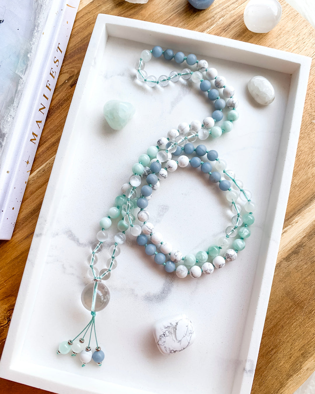 A hand-knotted mala necklace with Clear Quartz, Selenite, Green Moonstone, Angelite, White Howlite, and Rainbow Moonstone beads, displayed on a white marble tray, with a clear quartz pendant at the centre, and surrounded by small crystals.