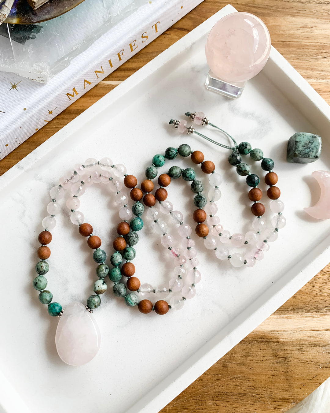 A hand-knotted mala necklace with beads of African Turquoise, Rose Quartz, and Sandalwood, and silver accents, with a Rose Quartz teardrop shaped pendant, displayed on a marble tray with small crystals.