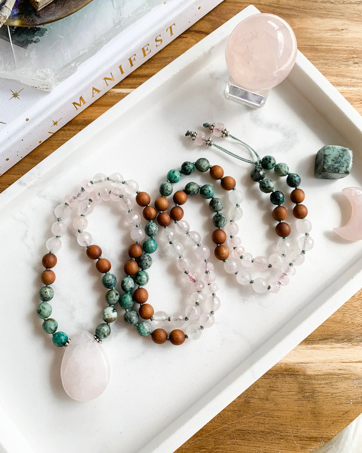 A hand-knotted mala necklace with beads of African Turquoise, Rose Quartz, and Sandalwood, and silver accents, with a Rose Quartz teardrop shaped pendant, displayed on a marble tray with small crystals.