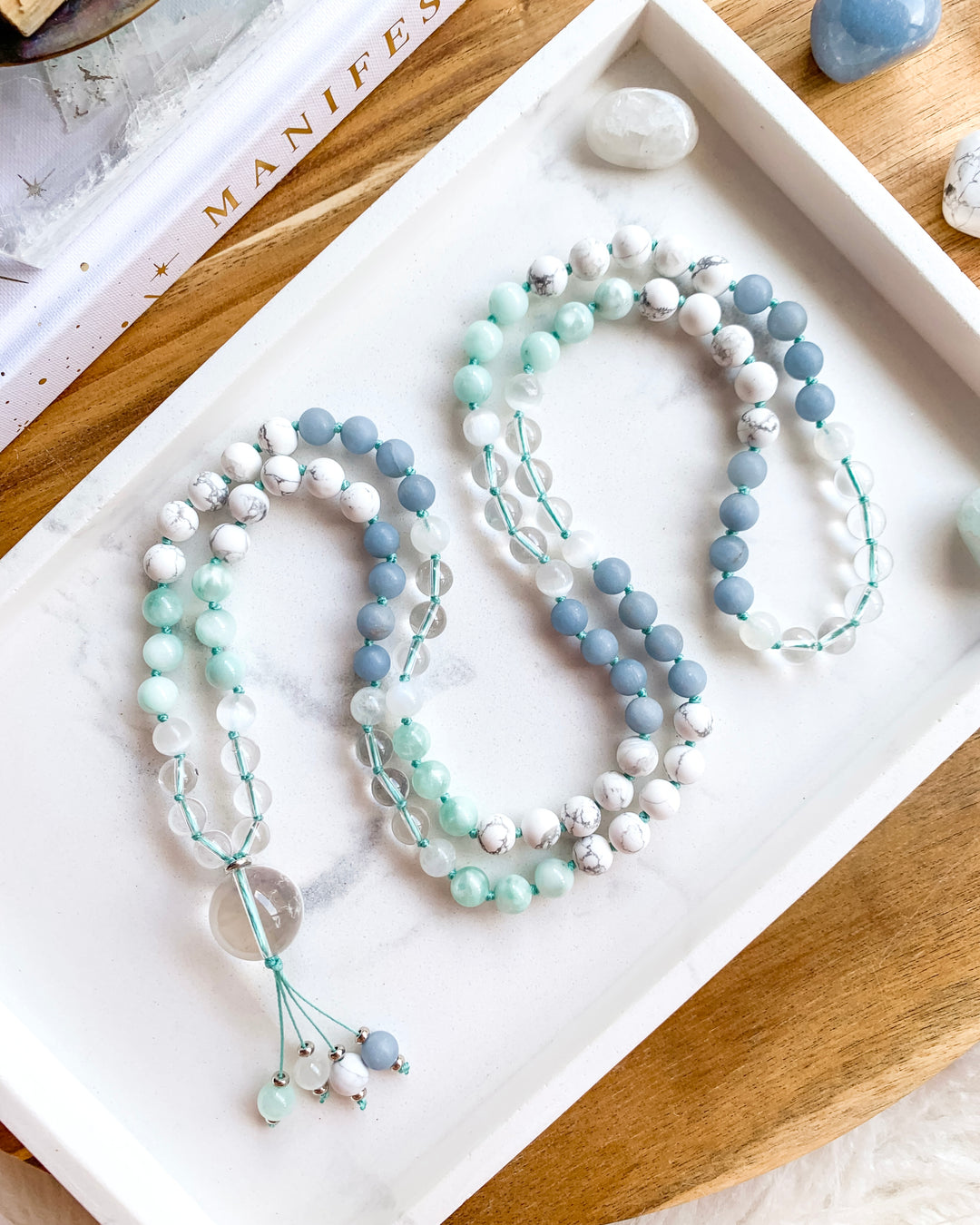 A hand-knotted mala necklace with Clear Quartz, Selenite, Green Moonstone, Angelite, White Howlite, and Rainbow Moonstone beads, displayed on a white marble tray, with a clear quartz pendant at the centre, and surrounded by small crystals.