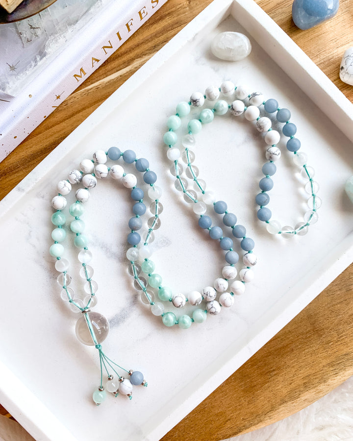 A hand-knotted mala necklace with Clear Quartz, Selenite, Green Moonstone, Angelite, White Howlite, and Rainbow Moonstone beads, displayed on a white marble tray, with a clear quartz pendant at the centre, and surrounded by small crystals.